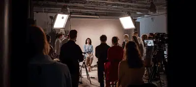 A woman sits on a stool in front of a backdrop, surrounded by a group of people and professional lighting equipment, suggesting filming in Moscow within a vibrant studio setting.