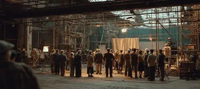 A group of people stands in a large, industrial-looking space with scaffolding and equipment, gathered around a curtain and camera setup—suggesting filming in Berlin on a warehouse-like film set.
