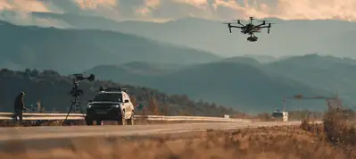 A large drone flies above a rural road with mountains in the background. A person stands beside a car fitted with a mounted camera, evoking the feel of filming in Berlin, all set under soft, warm light.