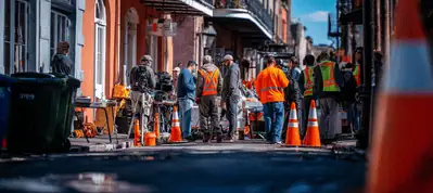 A group of construction workers in orange vests and helmets work on a city street in New Orleans, surrounded by orange traffic cones and buildings, under a blue sky—possibly filming in New Orleans.