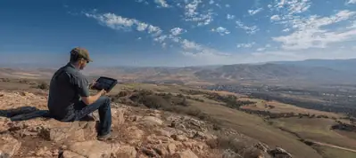 A person sits on a rocky hilltop using a laptop, overlooking a vast landscape of fields and hills—capturing the essence of filming in South Africa beneath a bright blue sky dotted with scattered clouds and a distant town.