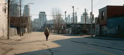 A person walks alone down an empty, cracked city street lined with old buildings and graffiti, filming in Austin, with modern skyscrapers visible in the distant skyline under a clear sky.