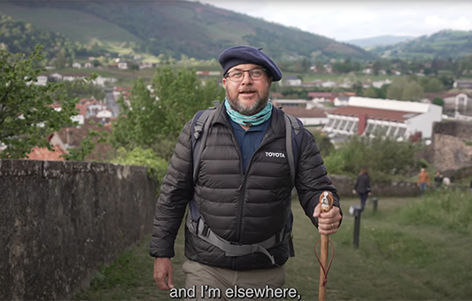 A man in a beret, glasses, and a puffy TOYOTA jacket stands on a path with a walking stick, green hills and a French town behind him. Subtitles read: and I’m elsewhere,.