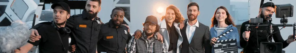 A diverse group of film crew members and actors, some in uniforms and others in business attire, stand together outdoors smiling, with a camera and clapperboard visible on set.