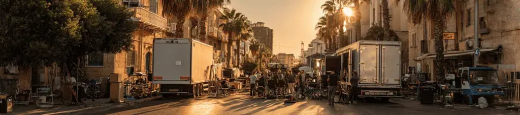 A film crew sets up equipment and trucks on a sunlit urban street lined with palm trees and old buildings while filming in Israel, as the setting sun casts a warm golden light over the scene.