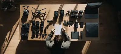 A person sits at a desk with a notepad, surrounded by neatly arranged cameras, lenses, a drone, a laptop, and photography equipment—everything needed for filming in Vienna—viewed from above in natural light.