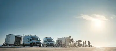 Film crew gathers around cameras and equipment near three white vans and trailers under a bright, clear sky on a wide, open outdoor set in Norway, with the sun shining—a perfect day for filming in Norway.