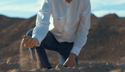 A person in a white shirt and jeans kneels on sandy ground in a desert landscape, letting sand fall through their fingers with mountains in the background—a striking scene perfect for a NEEDaFIXER Documentary Showreel.