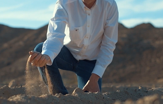 A person in a white shirt and jeans kneels on sandy ground in a desert landscape, letting sand fall through their fingers with mountains in the background—a striking scene perfect for a NEEDaFIXER Documentary Showreel.