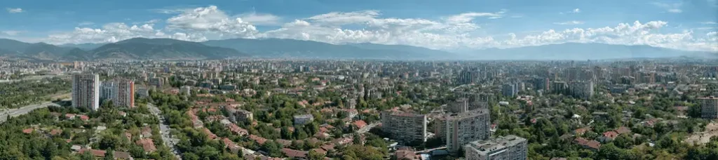 Panoramic view of Sofia with many buildings, green trees, and a distant mountain range under a blue sky with scattered clouds—perfect for filming in Sofia.