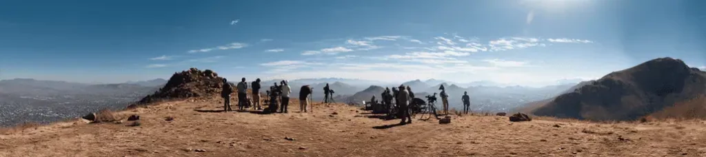 A group of people with cameras and equipment stand on a dry, rocky hilltop, filming in South Africa, overlooking a sprawling valley and distant mountains under a clear blue sky.