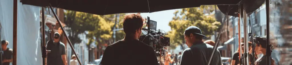 A film crew sets up a camera and equipment outdoors on a sunny day, filming in South Africa, with several people working under a canopy and trees and buildings visible in the background.
