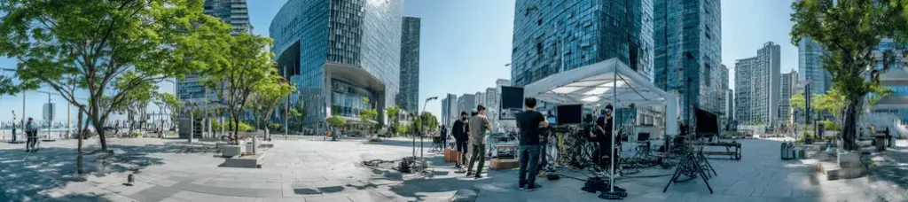 A group of people set up audio and camera equipment under a white tent in an urban plaza with modern glass buildings, trees, and bright sunlight, capturing the vibrant atmosphere while filming in South Korea.