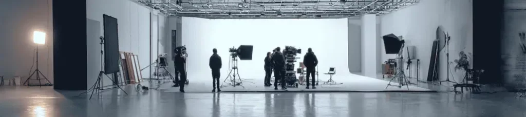 A film crew is filming in South Korea, setting up cameras, lights, and equipment in a spacious, brightly lit studio with a white backdrop. Several people stand and work around the filming area.