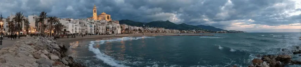 Panoramic view of a coastal town near Barcelona, perfect for filming, with a sandy beach, palm trees, historic church, and mountain backdrop beneath a dramatic cloudy sky at dusk as gentle waves touch the shore.