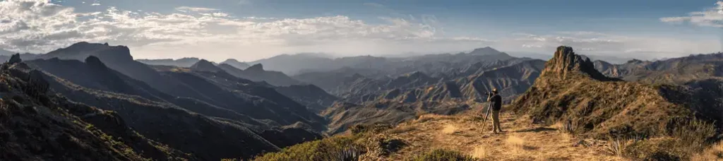 A panoramic view of a hiker standing on a dry, grassy hilltop overlooking a vast, rugged mountain landscape under a partly cloudy sky—ideal for filming in Vietnam, with warm sunlight and distant peaks fading into the horizon.