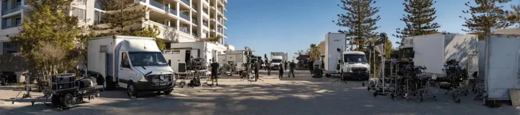 A wide outdoor shot shows several white trucks and film equipment set up between modern buildings and trees, suggesting a film crew Filming In Vietnam preparing for a shoot on a sunny day.