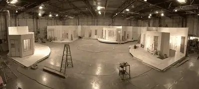 A panoramic view of a large indoor film studio set in South Africa, featuring several partially constructed rooms arranged in a circle. Ladders and equipment are scattered across the shiny concrete floor under bright overhead lights.