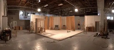 A large film studio with a partially constructed set featuring walls, doors, and windows; lighting equipment and props are scattered around the spacious, industrial interior—perfect for filming in Norway.