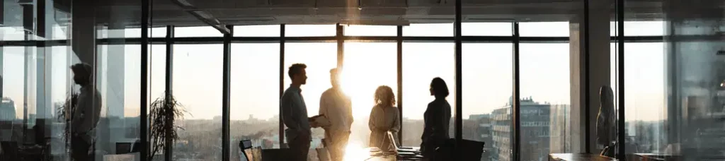 Five people stand and talk in a modern office with large floor-to-ceiling windows. Sunlight streams in, casting silhouettes and illuminating city buildings outside while filming in Sweden.
