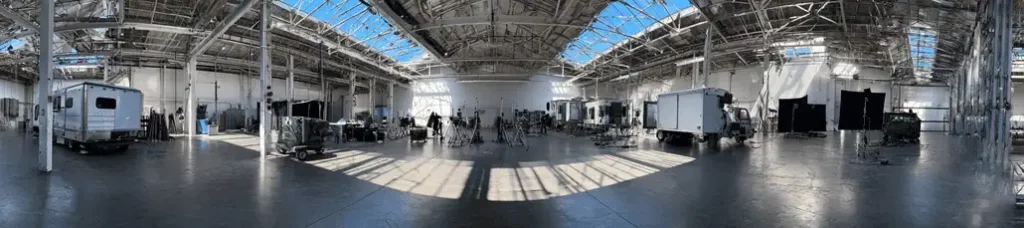 Wide panoramic view of a spacious, sunlit warehouse in Sweden with trucks, trailers, equipment, and crew members, suggesting a filming in Sweden setup under a high, skylit ceiling.