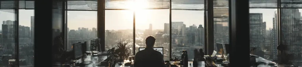 A person sits at a desk in a modern office with large windows, working on a computer while looking out at a city skyline at sunset, reflecting on filming in Sweden.