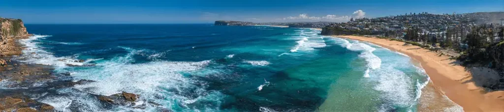 A panoramic view of a coastal beach, perfect for filming in Sydney, with turquoise waves, golden sand, rocky cliffs on the left, and a seaside town nestled among trees on the right under a clear blue sky.