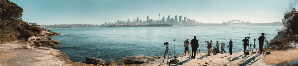 A panoramic view of photographers with tripods on a rocky shoreline, filming in Sydney as they capture the iconic skyline and Sydney Harbour Bridge across the water beneath a clear blue sky.