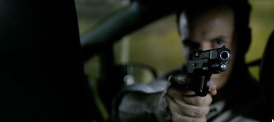 A person sitting inside a car aims a handgun directly at the camera, with their face partially obscured and out of focus in the background—a dramatic scene perfect for a video production company in Italy.