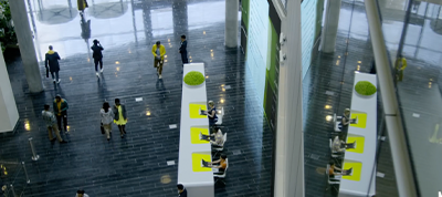 A modern building lobby in Italy with people walking and some seated at white desks with bright yellow accents; large windows and reflective floors create an open, bright atmosphere ideal for a video production company.