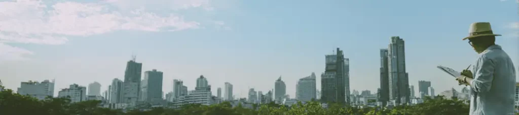 A person in a light shirt and hat stands in front of a city skyline with tall buildings under a blue sky, writing on a clipboard, with green trees in the foreground—perfect for filming in Vietnam.