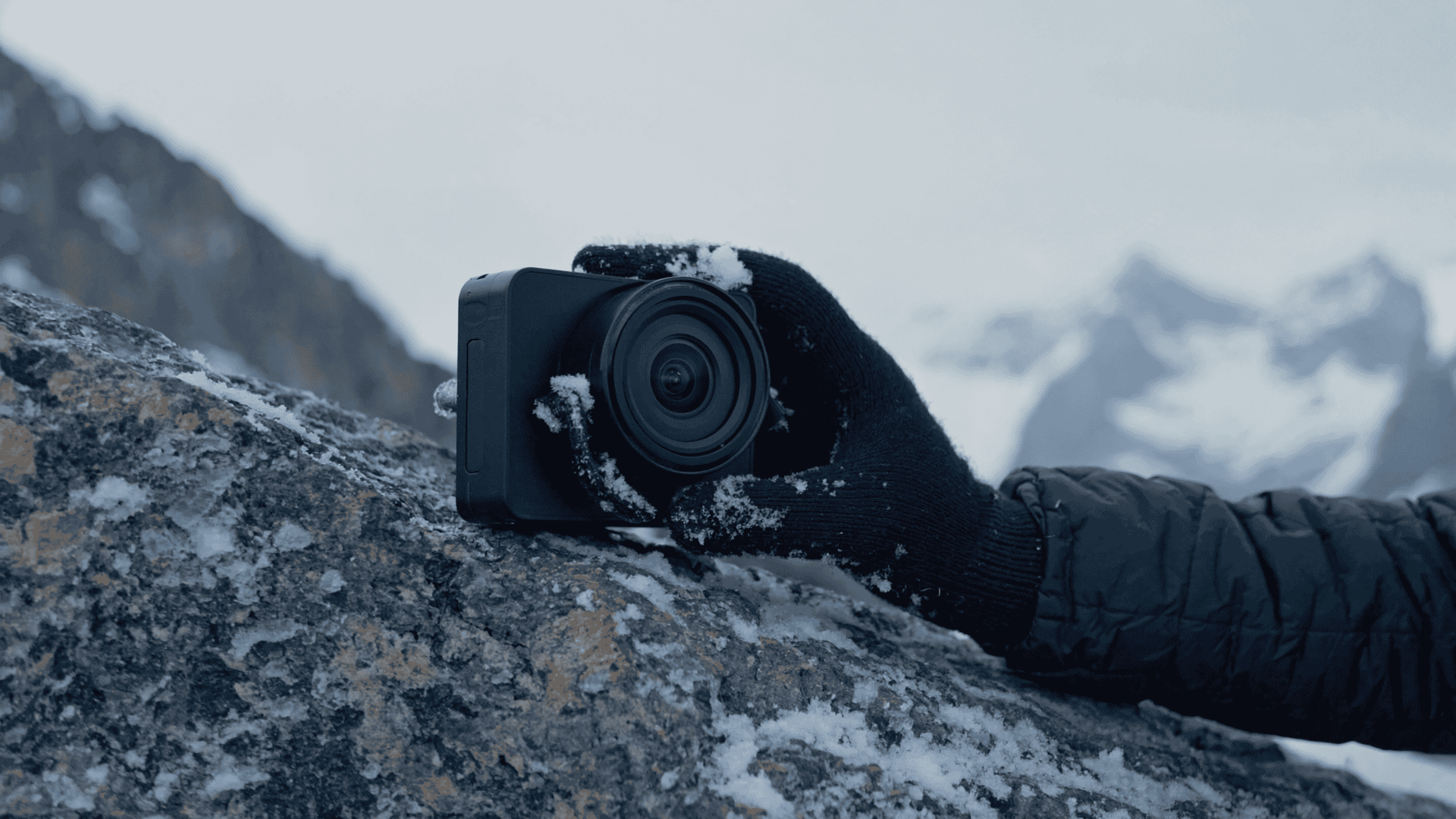 A gloved hand holding a camera covered with snow rests on a snow-dusted rock, capturing the essence of extreme environment photography with snowy mountains blurred in the background.