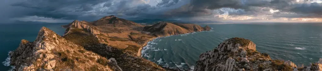 A panoramic view of a rugged coastline near Tirana reveals rocky hills, rolling terrain, and a curved sandy beach meeting the wavy sea under dramatic, cloudy skies—an inspiring location for filming in Tirana.