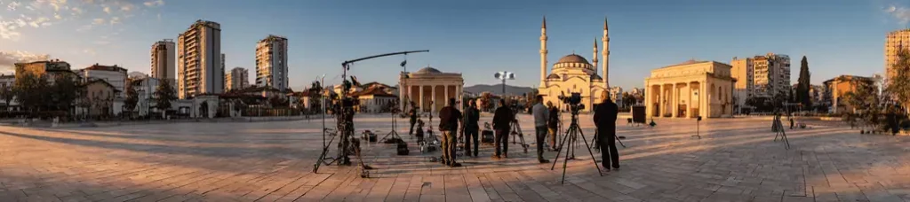 A wide plaza with a film crew setting up cameras and equipment for filming in Tirana, surrounded by modern buildings, a mosque with twin minarets, and neoclassical structures at sunset.