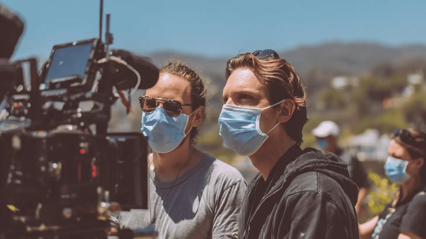 Two people wearing face masks stand outdoors in front of a professional video camera, working on a film set. Amid Covid-19, they follow safety protocols while filming during Covid at scenic film locations with clear skies and blurred greenery behind them.