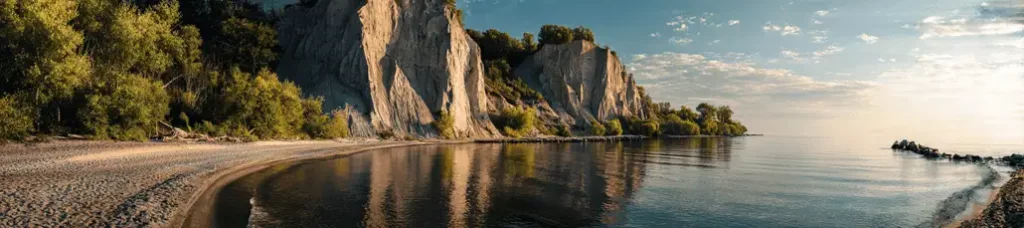 Panoramic view of a calm lake bordered by sandy shore, lush green trees, and tall, rocky cliffs under a partly cloudy sky during golden hour—perfect scenery reminiscent of filming in Toronto.
