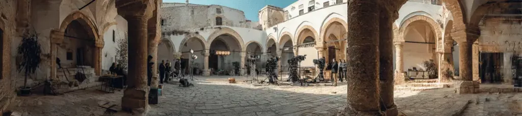 A film crew sets up cameras and equipment in a historic stone courtyard with arches and columns under a clear blue sky, preparing for a shoot and capturing the unique atmosphere of Filming In Tunisia.