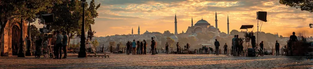 People stand and walk on a cobblestone square at sunset, with camera equipment set up for filming in Turkey. In the background, Istanbul’s skyline, including the Hagia Sophia and minarets, is silhouetted against a vibrant sky.