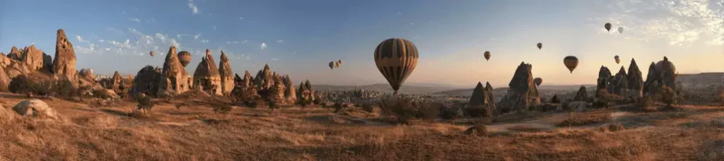 Panoramic view of a rocky landscape at sunrise with several hot air balloons floating in the sky over Cappadocia, Turkey. Dry grass and unique rock formations are perfect for filming in Turkey.