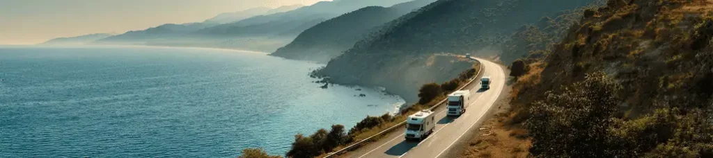 Three RVs drive along a winding coastal road bordered by the sea and mountains under soft sunlight—a perfect scene for filming in Turkey.