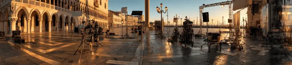 A panoramic view of filming in Venice at sunrise, with cameras, equipment, and lighting arranged on wet cobblestones near ornate old buildings and classic lamp posts in a historic European square.