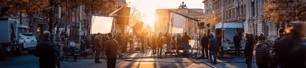 A film crew works on a city street at sunset, surrounded by equipment, lights, and camera rigs while filming in Berlin, with historic buildings and an archway visible in the background.