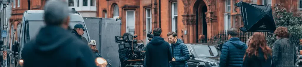 A film crew sets up cameras and equipment on a city street with historic brick buildings in the background, filming in Birmingham as several people stand nearby, preparing for a shoot.