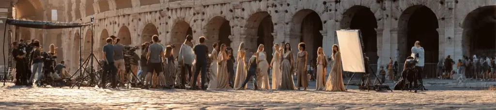 A film crew and several actors in ancient Roman-style costumes stand in front of the Colosseum, surrounded by lighting equipment and cameras, filming in Rome during a movie shoot at sunset.