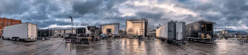 A wide view of a film production set outdoors in Wellington, featuring trailers, lighting equipment, and large light panels on a wet concrete lot under a cloudy sky.