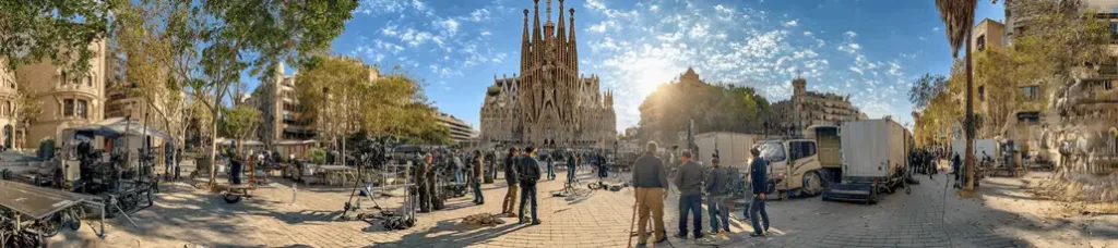 A group of people and filming equipment are set up in a sunny plaza near the Sagrada Familia, capturing the essence of filming in Barcelona with the iconic basilica and cityscape visible under a blue sky dotted with clouds.