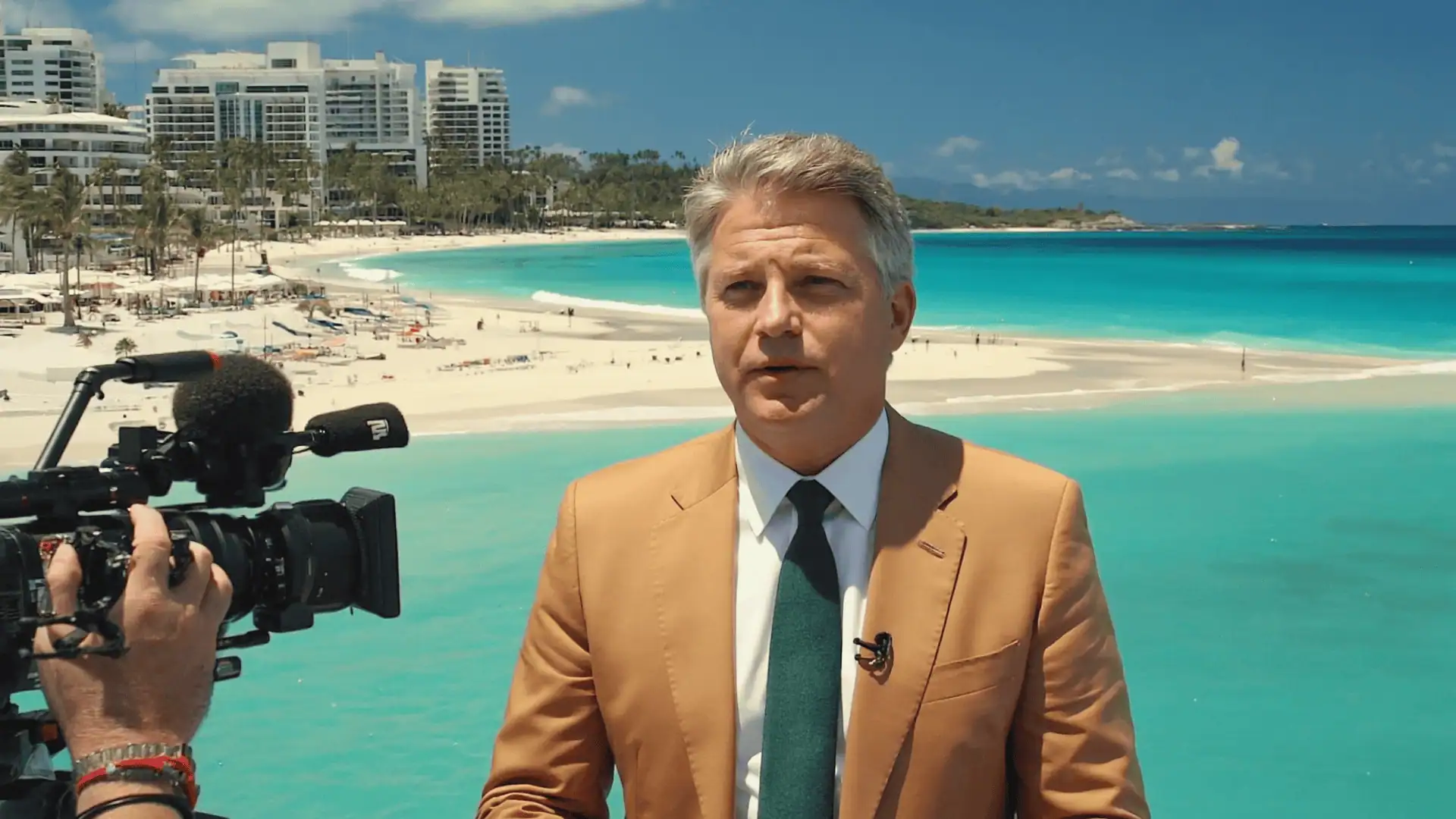A man in a tan suit and green tie stands in front of a bright blue ocean and sandy beach, speaking on camera for a business video. Modern high-rise buildings and palm trees are visible in the background under a sunny sky.