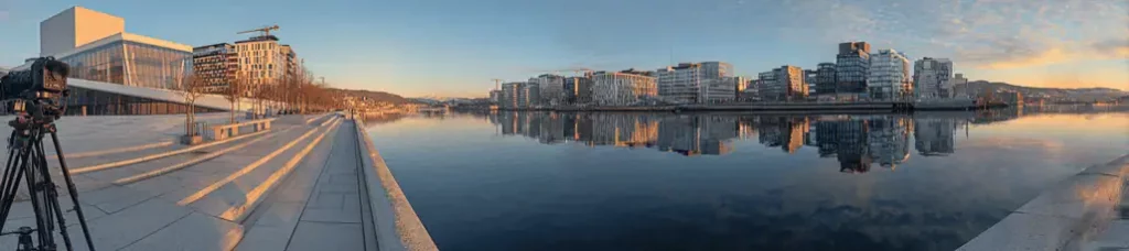 Panoramic view of a modern city waterfront at sunrise or sunset in Oslo, showing glass buildings reflected in calm water, concrete steps, and a camera on a tripod on the left—perfect for filming in Oslo under mostly clear skies with some clouds.