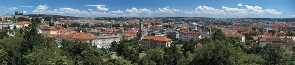 Panoramic view of a European city with red-roofed buildings, church spires, and lush green trees under a blue sky with scattered clouds—perfect for filming in Prague.