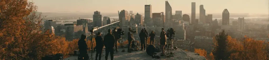 A group of people with cameras and tripods stand on a hill filming in Montreal, overlooking the city skyline at sunrise or sunset, with tall buildings and autumn trees in the foreground.